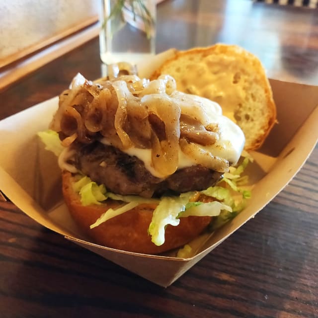 Red Pepper Harissa Burger featuring a grilled patty topped with caramelized onions and creamy sauce on a toasted bun, served with lettuce in a takeout box, highlighting the unique flavor profile discussed in the review.